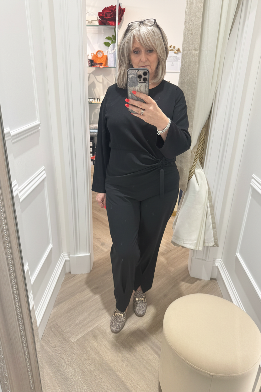 A woman takes a mirror selfie in a fitting room or boutique, wearing a black long-sleeve top and black pants. The room has white paneled walls, a light wood floor, and a cream-colored stool in the foreground.